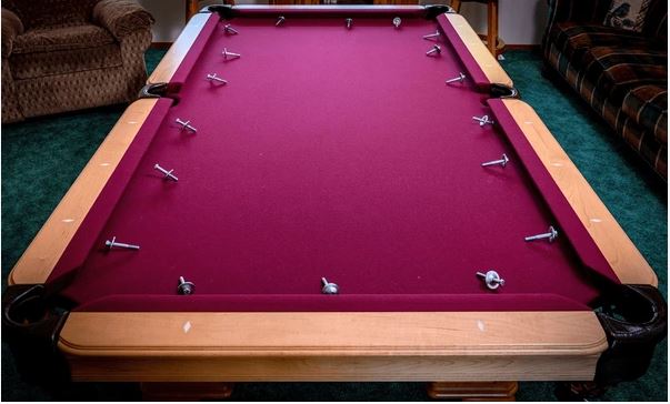 The image shows a burgundy-colored pool table with wooden rails and several white cue balls scattered on the felt surface, situated in what appears to be a basement or recreation room with carpeting and furniture visible in the background.Retry
