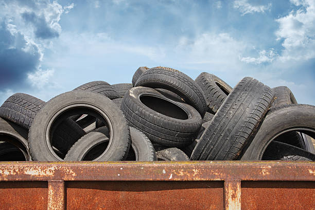 The image shows a large pile of old, used tires stacked in what appears to be a rusty metal container or dumpster against a cloudy blue sky.Retry